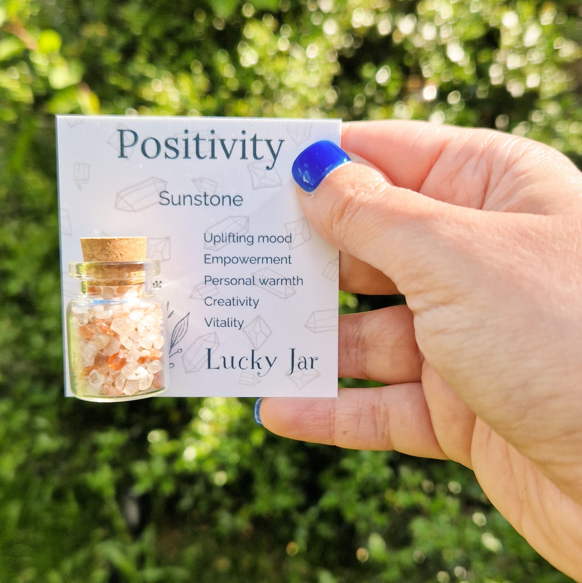 The image shows a hand with blue nail polish holding a card labelled "Positivity" featuring a small glass jar filled with sunstone crystal chips. The card lists the benefits of Sunstone. The jar has a cork stopper and is displayed against a blurred outdoor background with greenery, enhancing the natural and calming aesthetic of the product. The text "Lucky Jar" is prominently displayed at the bottom of the card, indicating the product's branding.