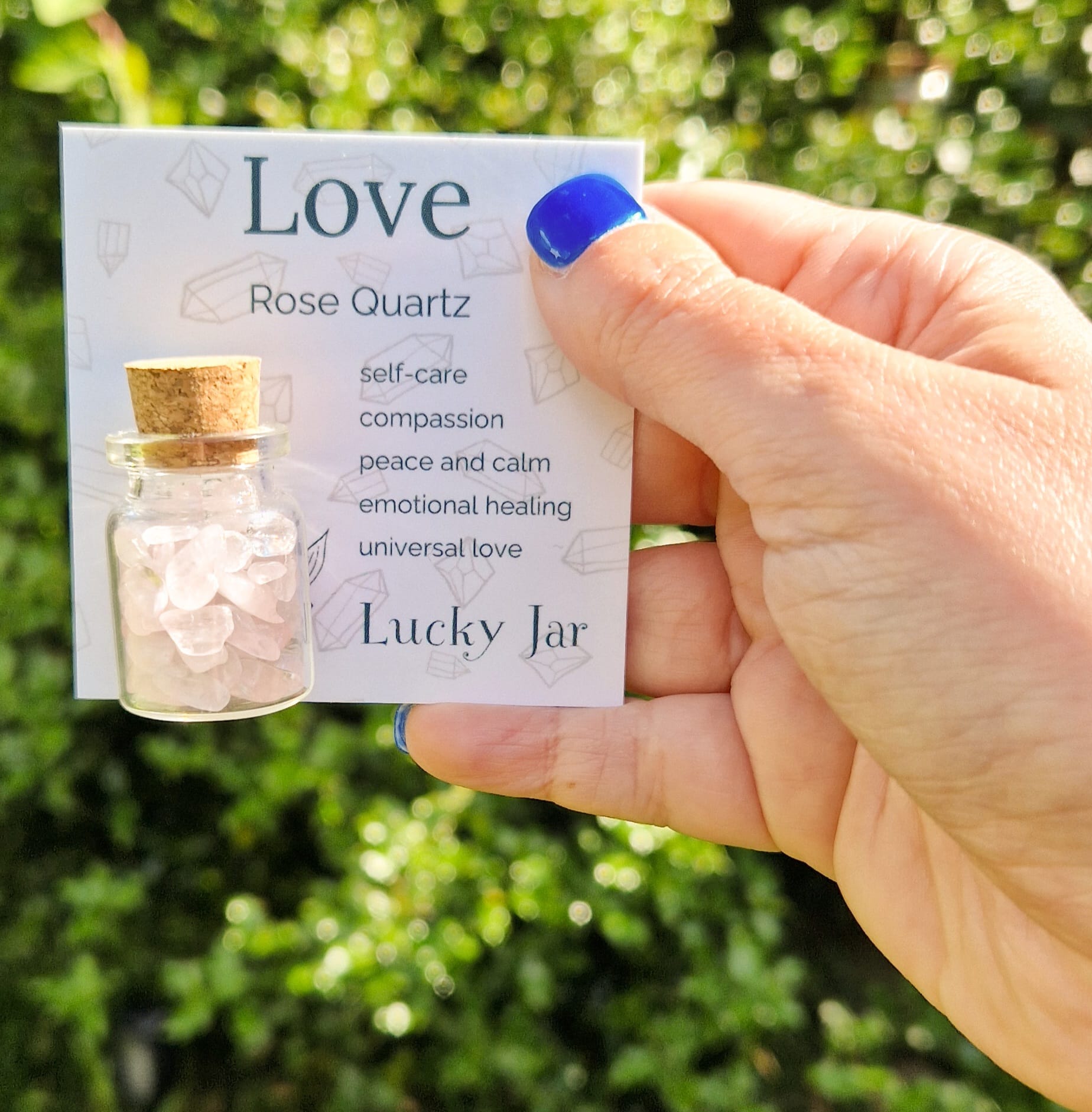 The image shows a hand with blue nail polish holding a card labelled "Love" featuring a small glass jar filled with rose quartz crystal chips. The card lists the benefits of Rose Quartz. The jar has a cork stopper and is displayed against a blurred outdoor background with greenery, enhancing the natural and calming aesthetic of the product. The text "Lucky Jar" is prominently displayed at the bottom of the card, indicating the product's branding.