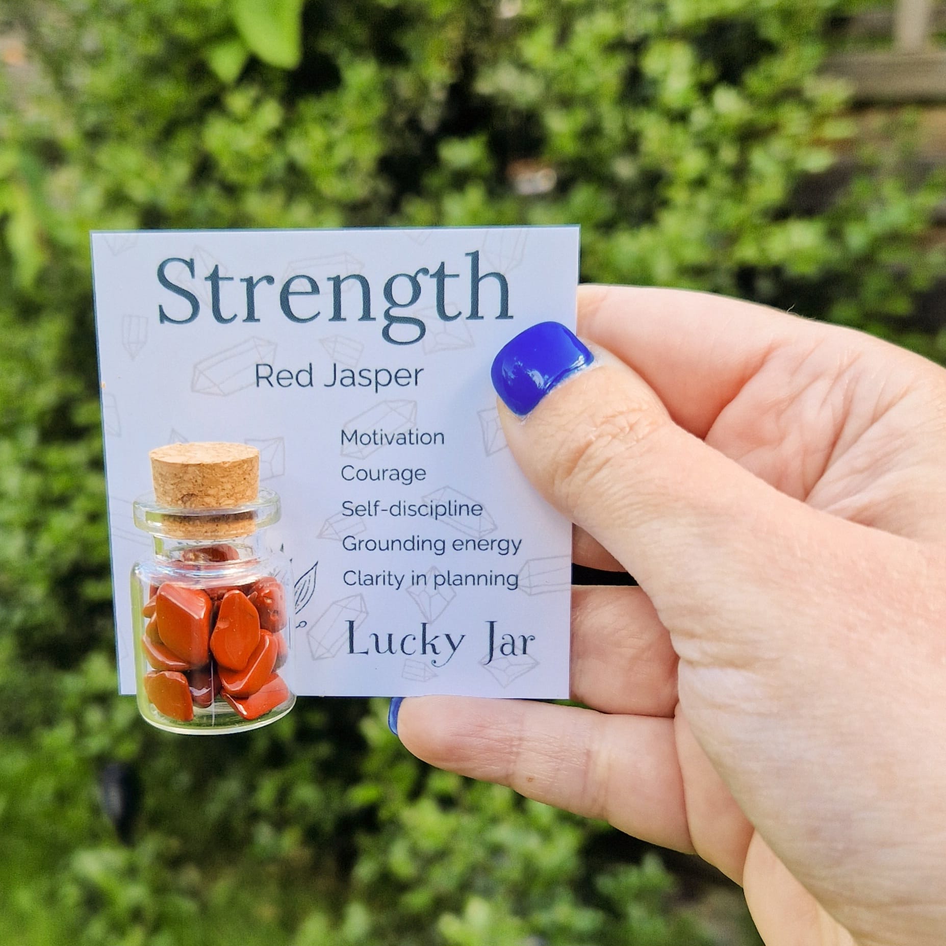 The image shows a hand with blue nail polish holding a card labelled "Strength" featuring a small glass jar filled with red jasper crystal chips. The card lists the benefits of Red Jasper.  The jar has a cork stopper and is displayed against a blurred outdoor background with greenery, enhancing the natural and calming aesthetic of the product. The text "Lucky Jar" is prominently displayed at the bottom of the card, indicating the product's branding.
