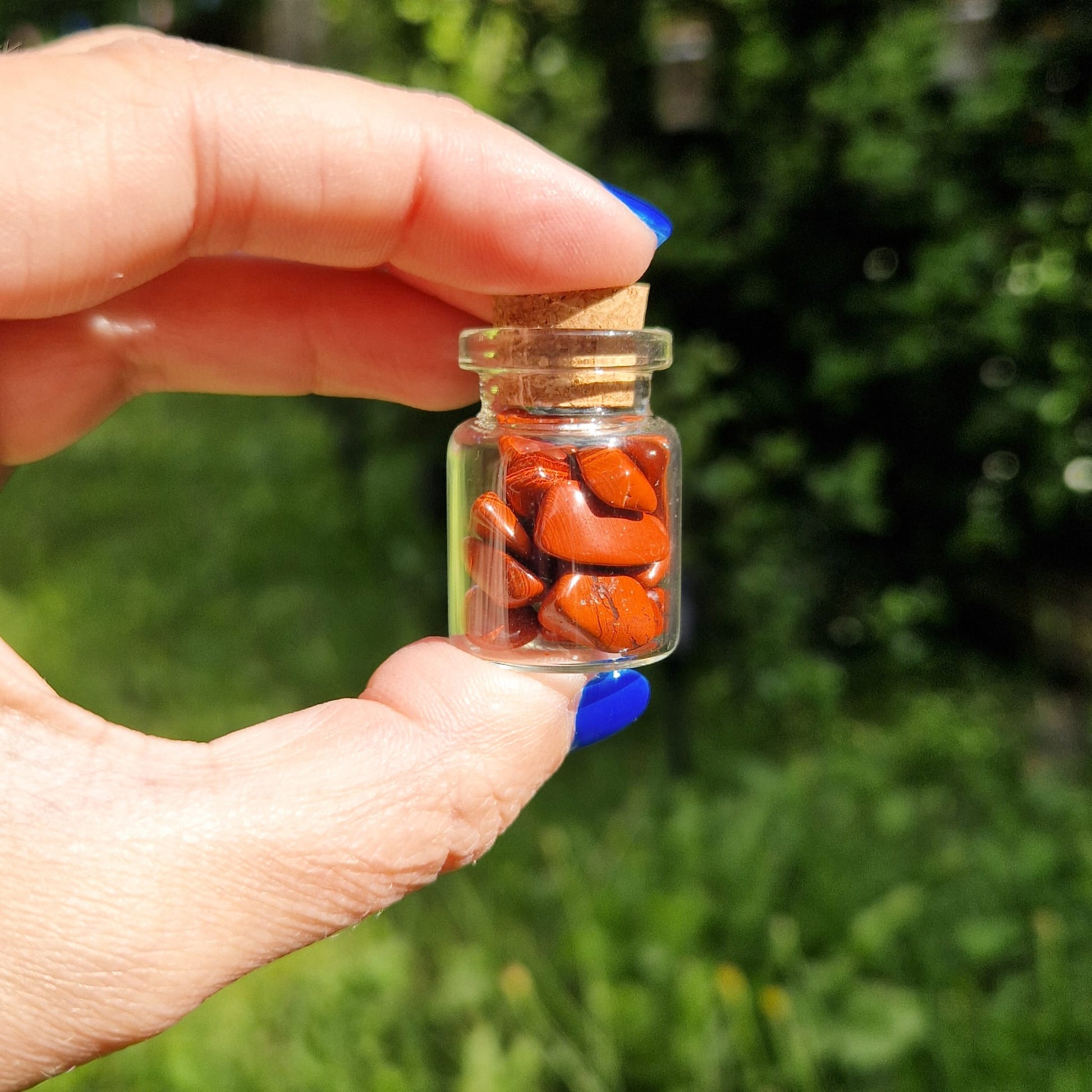 Small glass jar held between a thumb and forefinger, filled with red jasper crystal chips. The jar has a cork stopper and is being held outdoors, with greenery blurred in the background. The person holding the jar has blue nail polish.