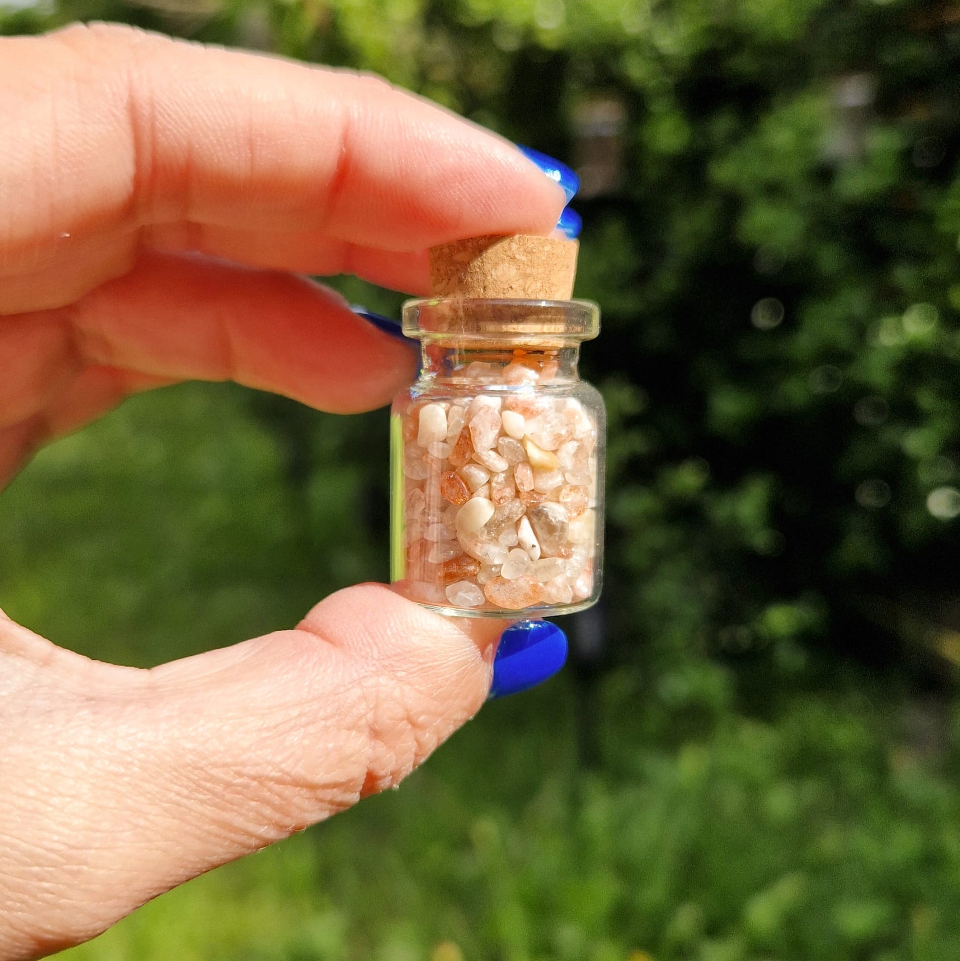 Small glass jar held between a thumb and forefinger, filled with sunstone crystal chips. The jar has a cork stopper and is being held outdoors, with greenery blurred in the background. The person holding the jar has blue nail polish.