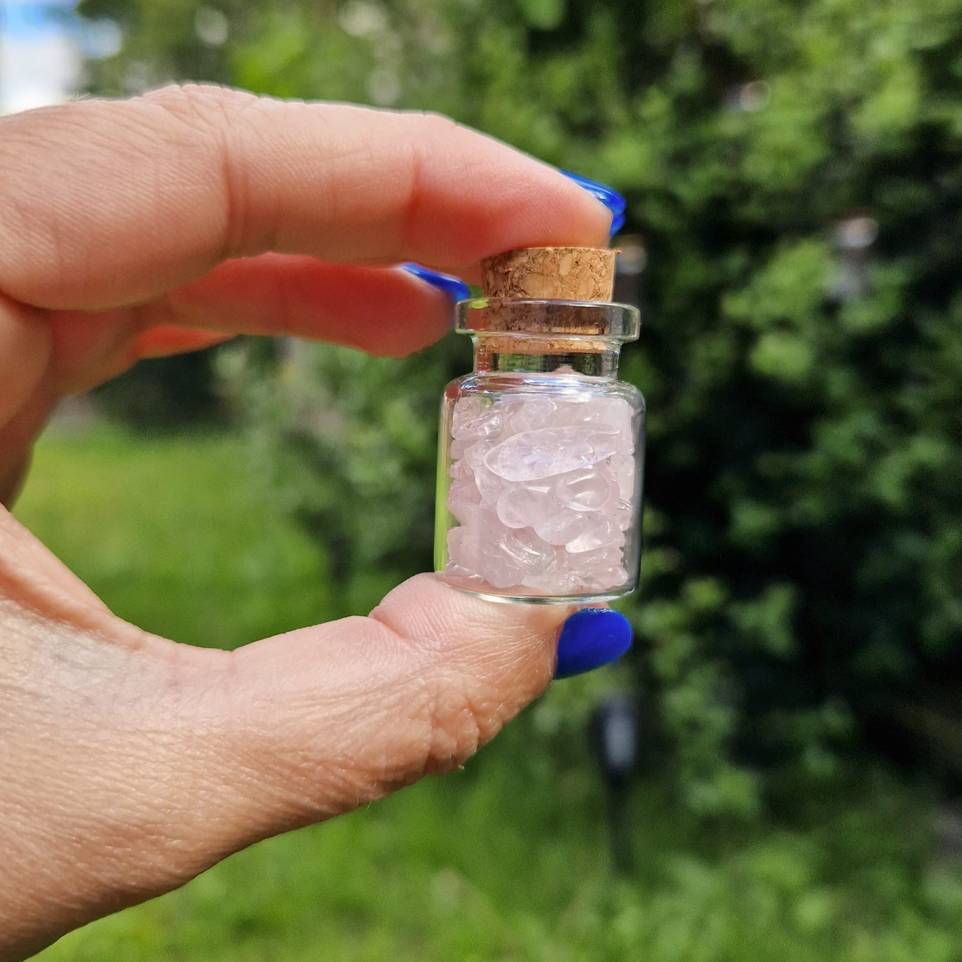 Small glass jar held between a thumb and forefinger, filled with rose quartz crystal chips. The jar has a cork stopper and is being held outdoors, with greenery blurred in the background. The person holding the jar has blue nail polish.