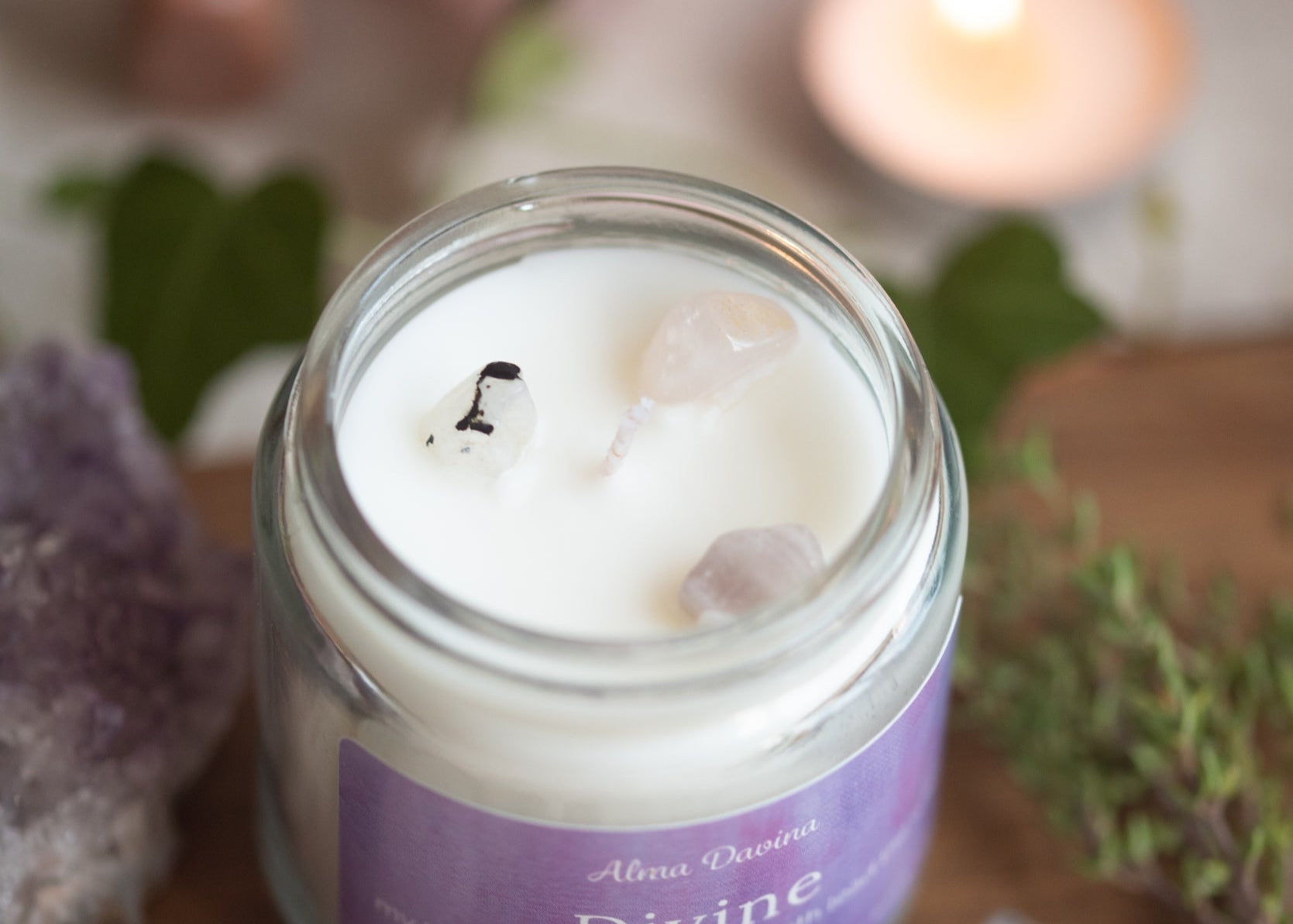 Candle in a jar labeled 'Divine' surrounded by crystals and plants on a wooden surface.